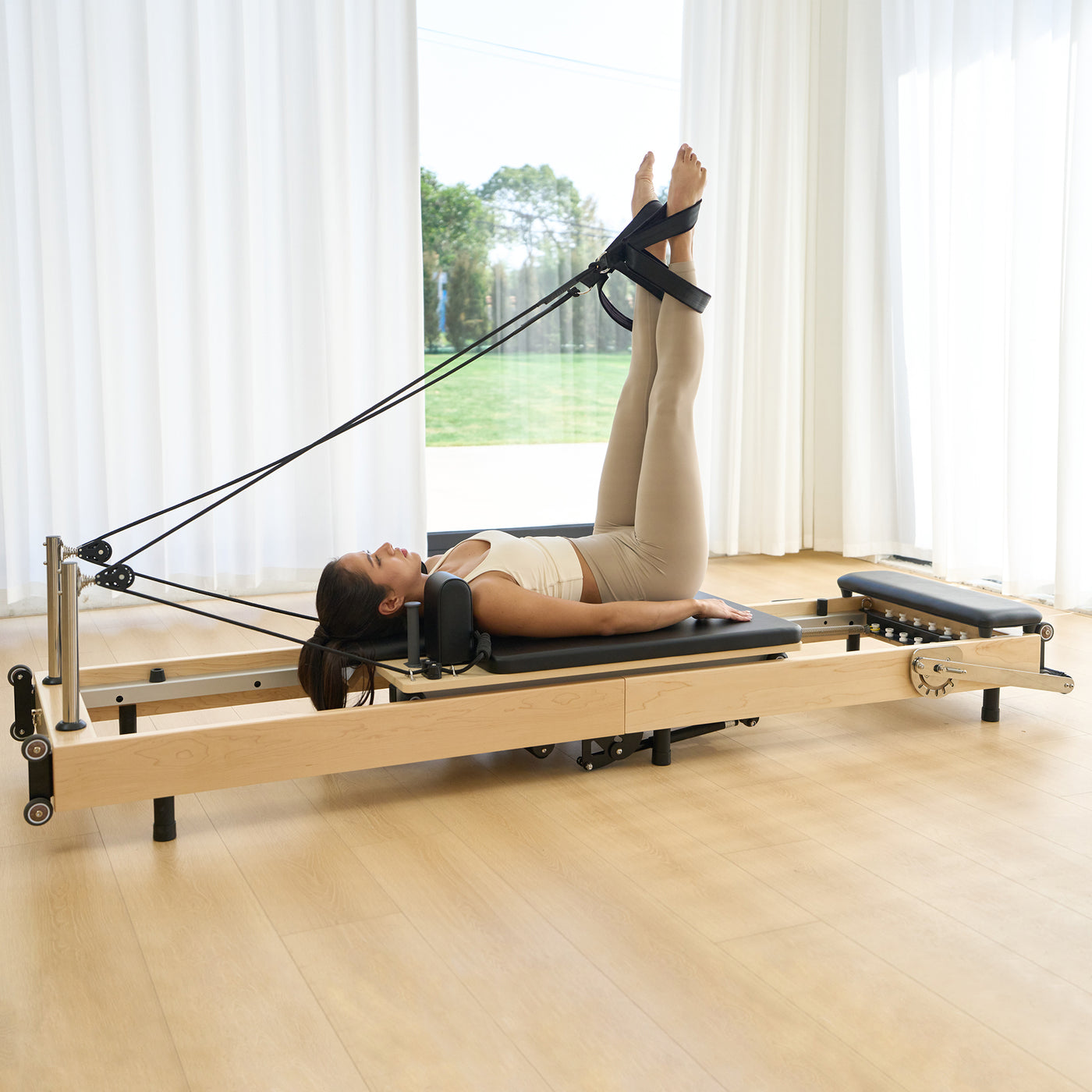 A woman in beige activewear lies on a wooden Pilates reformer machine with black straps and foot restraints, in a bright room with white curtains and a window.