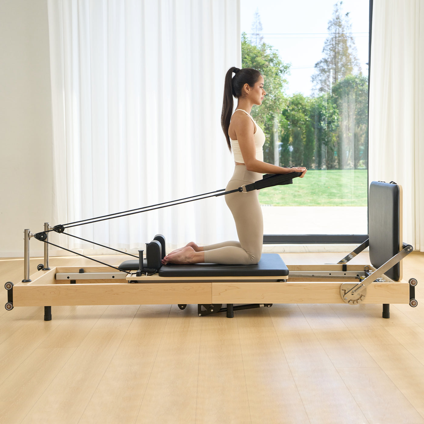 Woman using a Pilates reformer machine in a bright room with large windows.