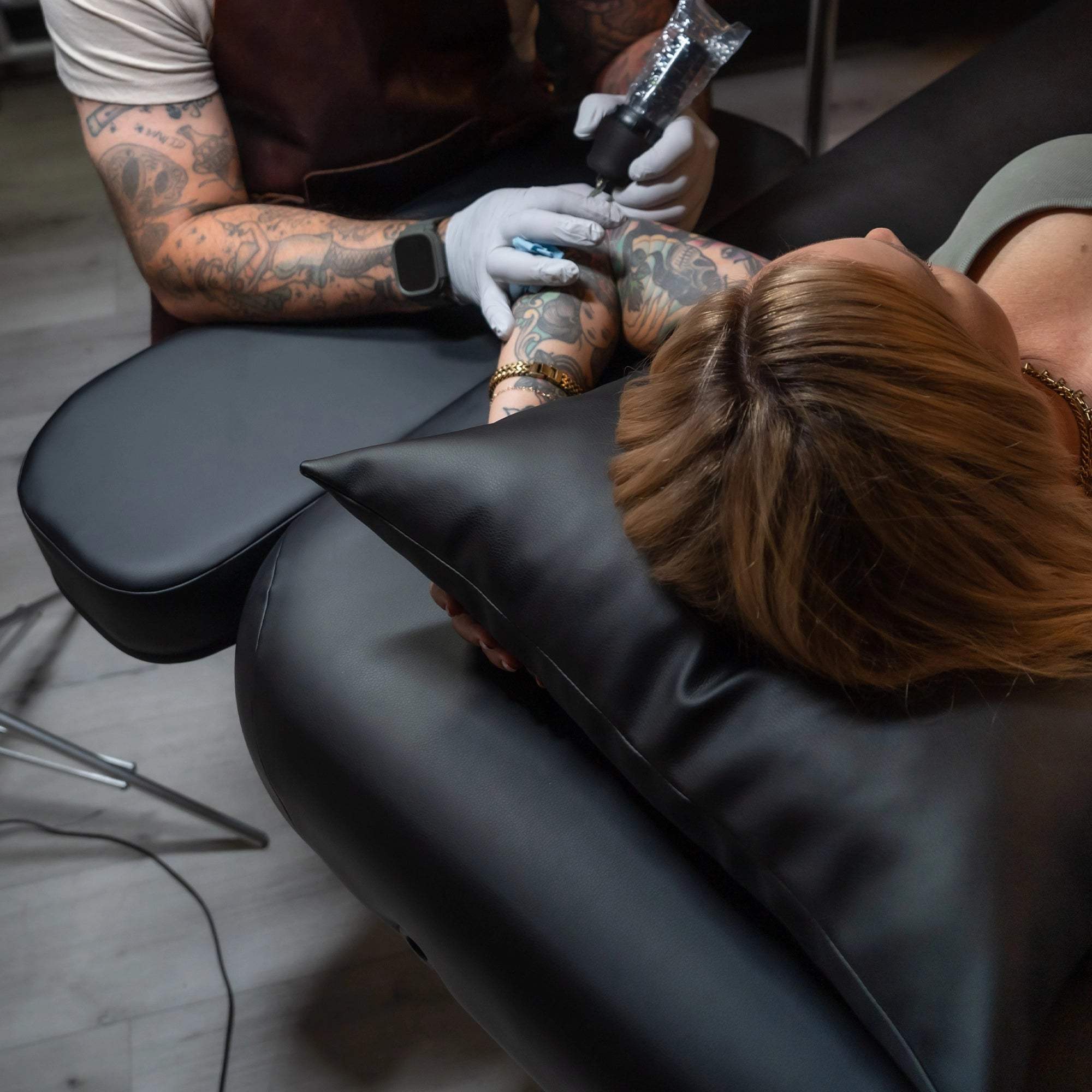 A tattoo artist with tattooed arms works on a client's forearm while the client reclines on a black Hydra table in a studio setting.