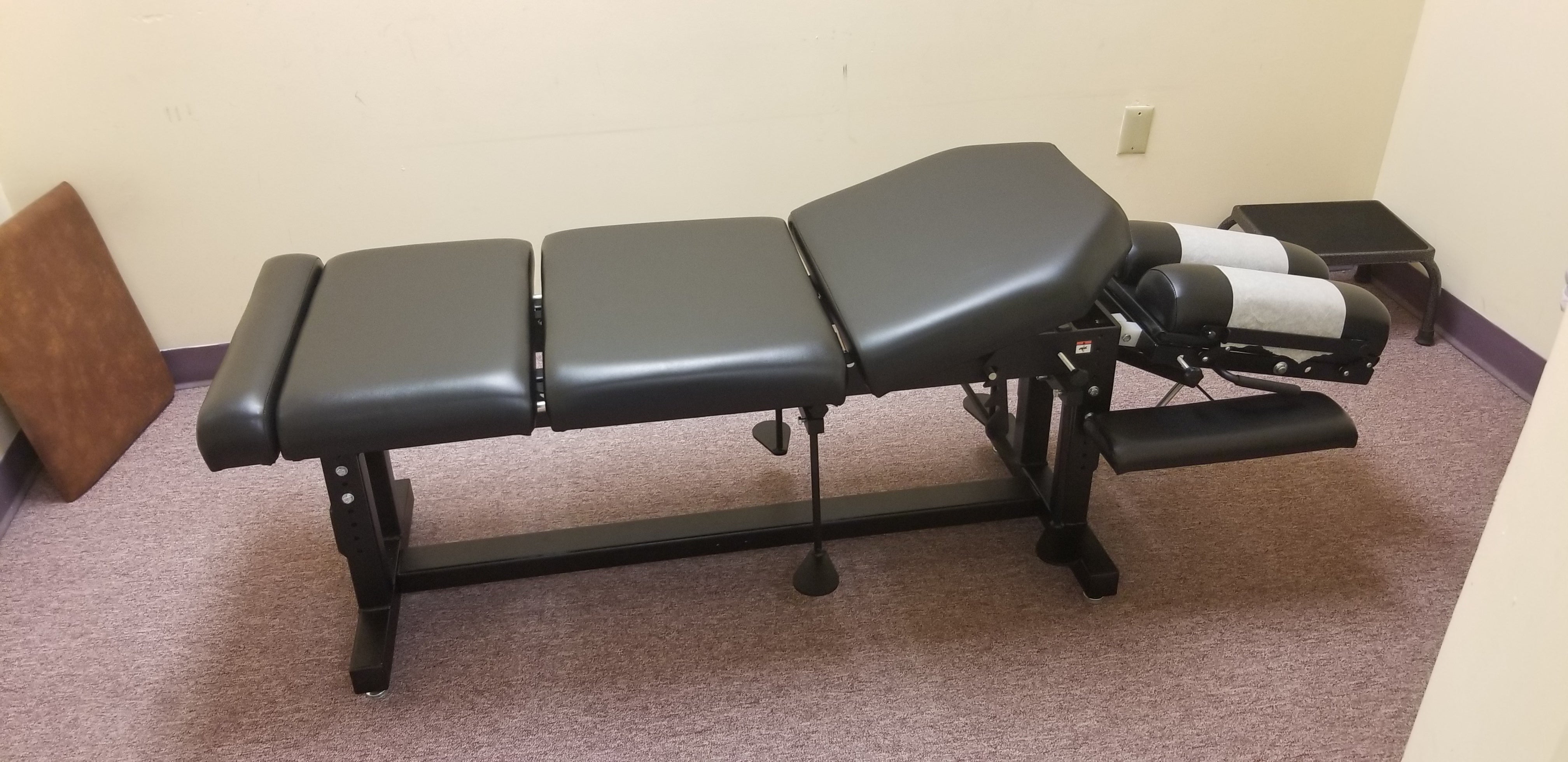 PHS METAL Max Drop Treatment Table by PHS Chiropractic in a small, carpeted, beige-walled room.