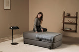 Woman sitting on a gray Ceragem S4 Massage Lounger in a room with a shelf and table.