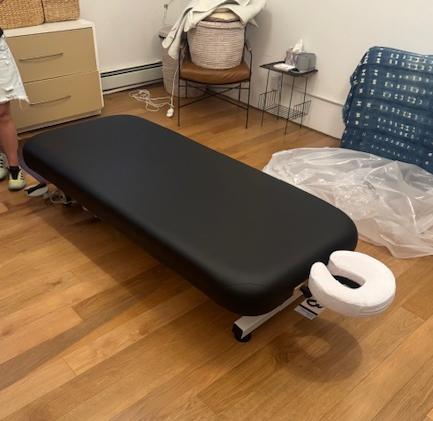 The EarthLite ELLORA LIFT black massage table with face cradle is in a wood-floored room.