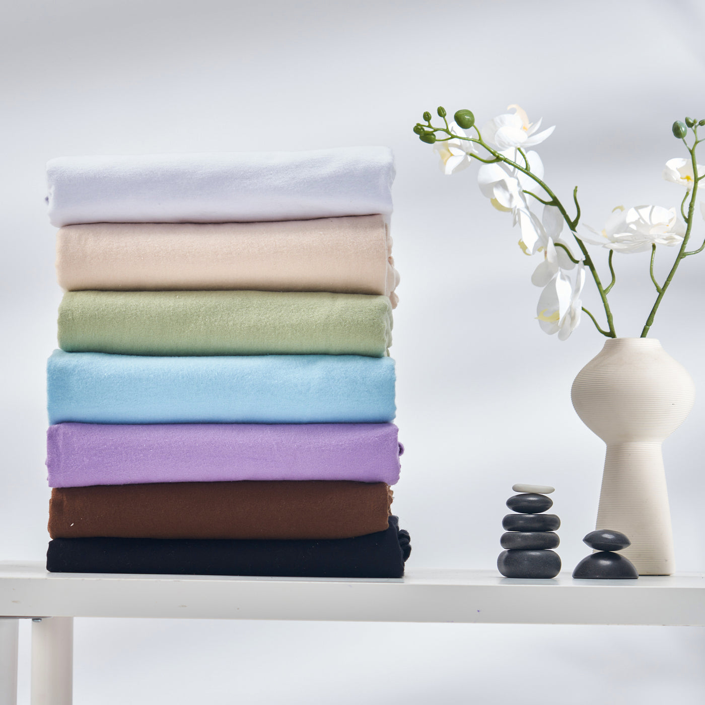Stack of folded Master Massage Cotton Flannel Sheets Set (3 Piece Set)  in various colors on a white surface with a vase of flowers and stones.