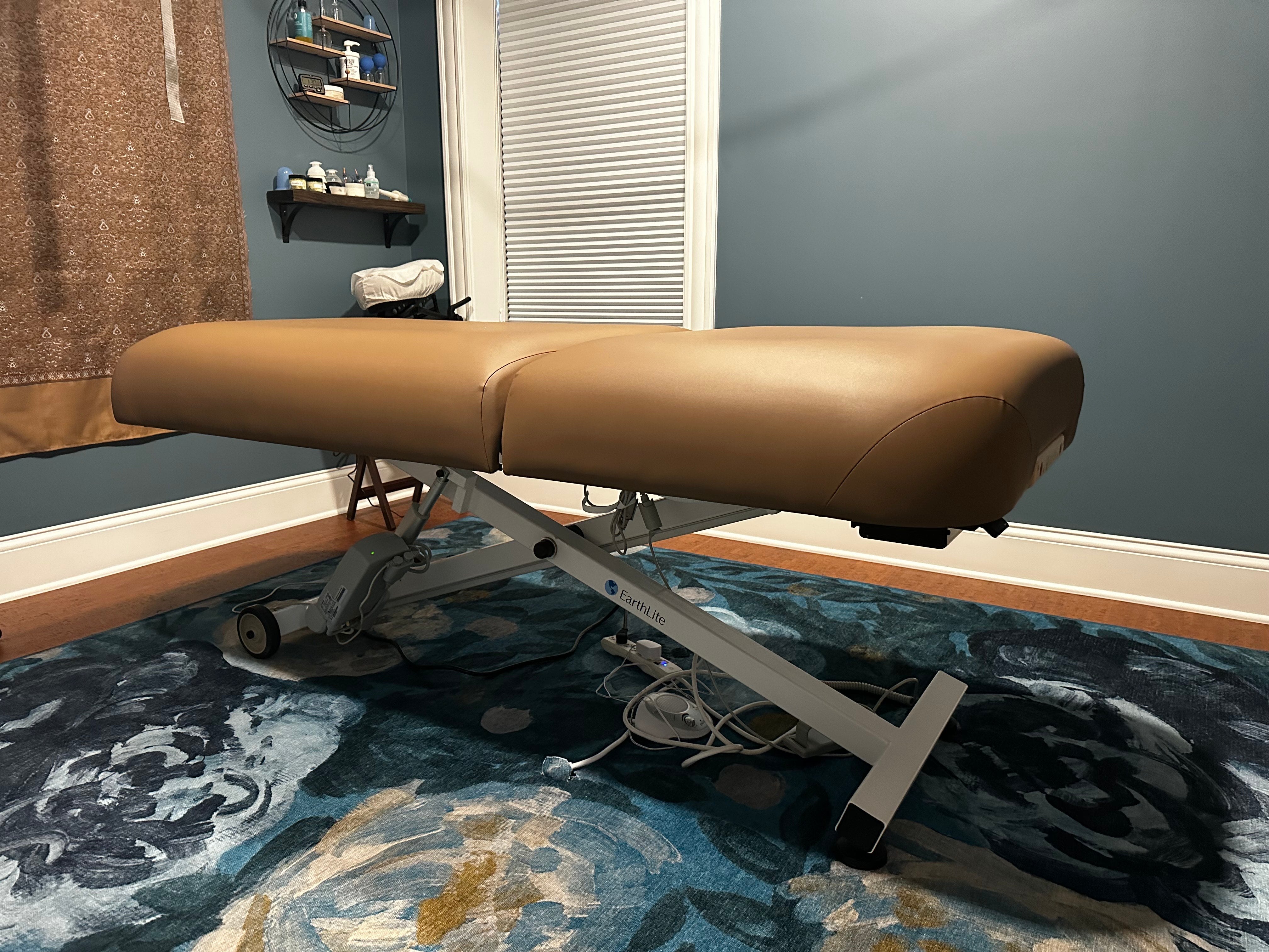 An EarthLite ELLORA TILT massage table sits in a dim room with blue walls and a patterned rug.