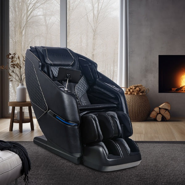 Black massage chair in a modern living room with large windows and a fireplace.