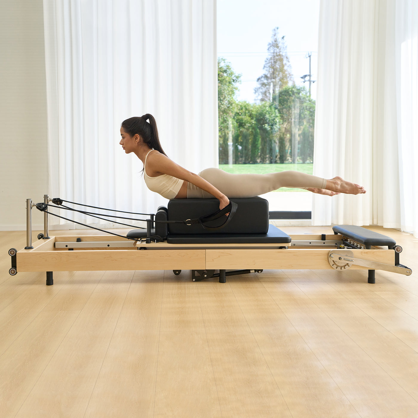 Woman using a Pilates reformer machine in a bright room with large windows.