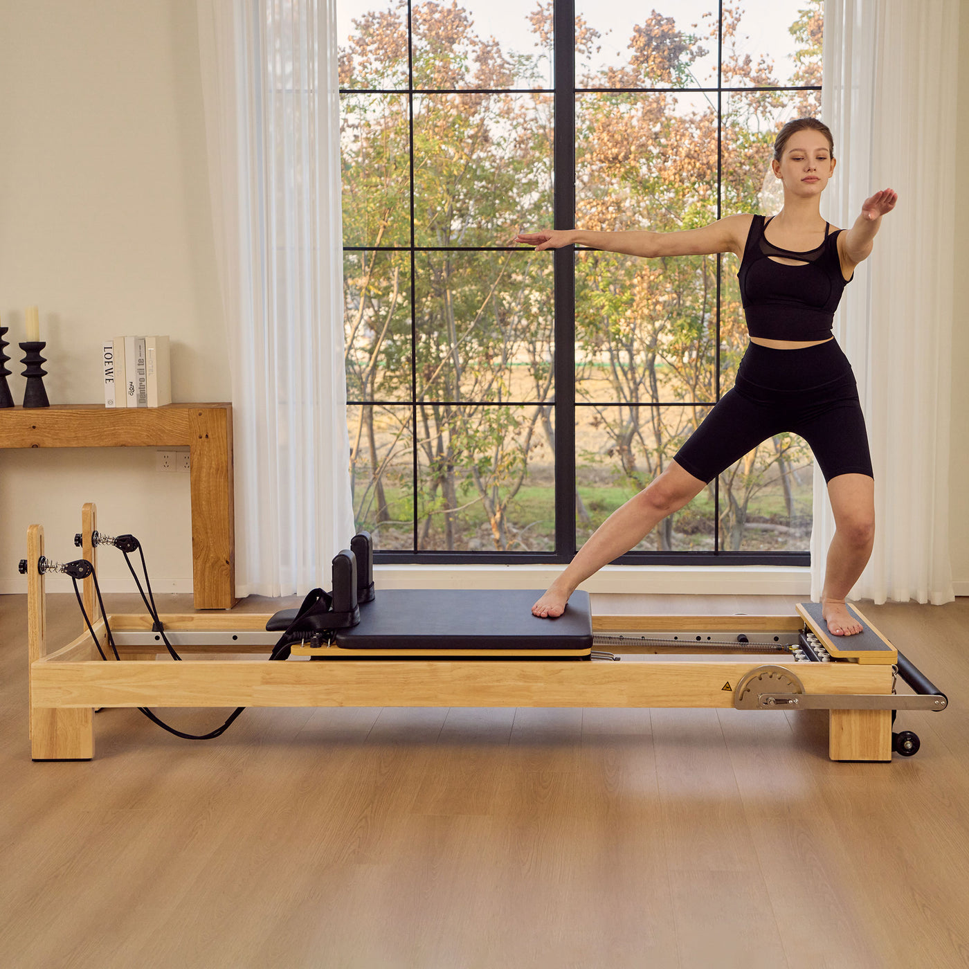 A woman in black activewear performs a Pilates exercise on Master Massage StudioSolid Stationary Pilates Reformer in a bright room with large windows and wood floors.
