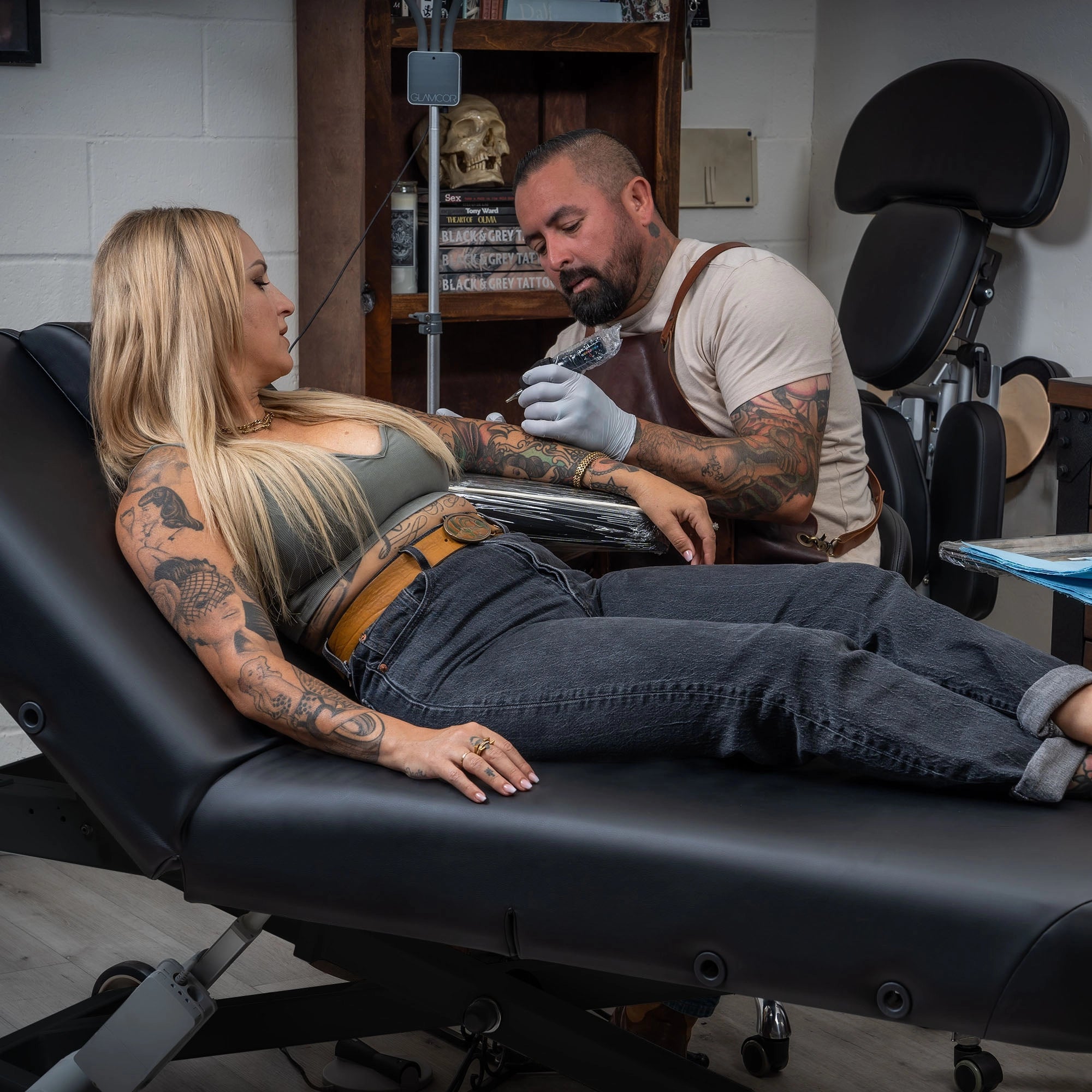 A tattoo artist works on a client with long blonde hair and visible arm tattoos, seated on a black Hydra tattoo table inside a tattoo studio.